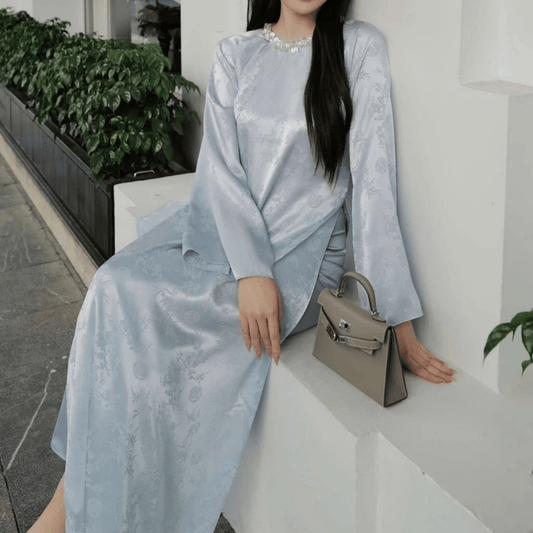 Woman in a light blue outfit holding a beige handbag on a white surface with greenery in the background