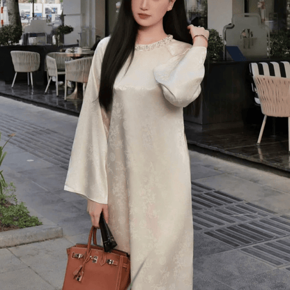 Woman in a long beige dress holding a brown handbag on a patio.